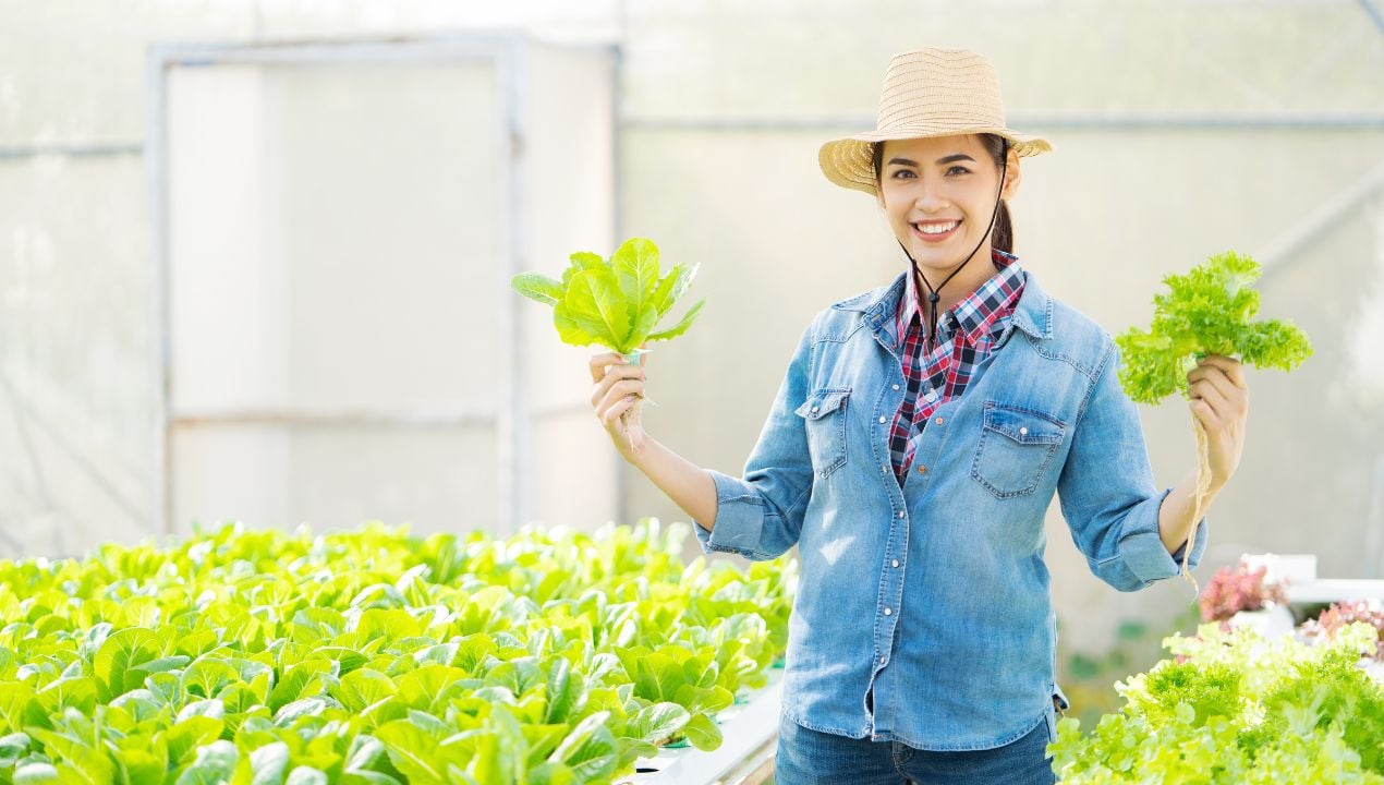 Woman inside a greenhouse holding up two different types of leafy greens standing in between the growing tables